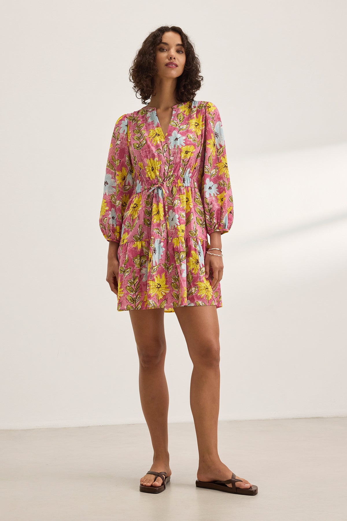 A person in the SHARLENE DRESS by Velvet by Graham & Spencer, featuring a pink floral pattern, a v-neckline, and a smocked elastic waist, stands in a well-lit indoor space, paired with flat sandals.-38663136772289