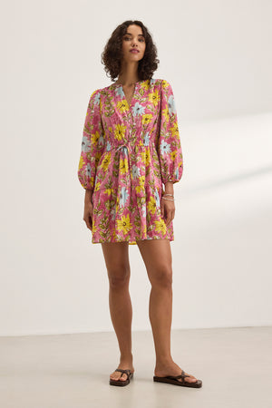 A person in the SHARLENE DRESS by Velvet by Graham & Spencer, featuring a pink floral pattern, a v-neckline, and a smocked elastic waist, stands in a well-lit indoor space, paired with flat sandals.