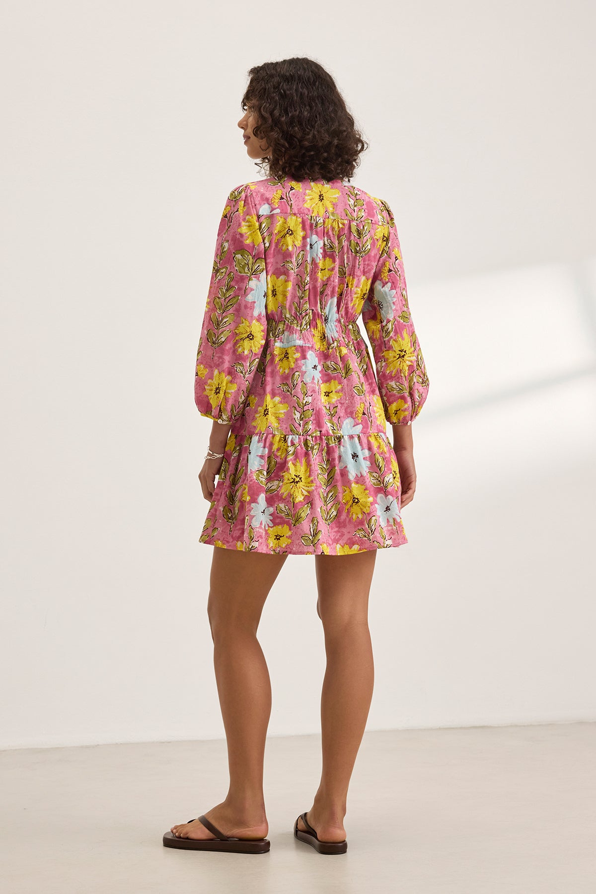   A woman stands with her back to the camera in the Velvet by Graham & Spencer SHARLENE DRESS, a pink floral mini dress featuring a smocked elastic waist, paired with sandals against a plain backdrop. 