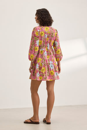 A woman stands with her back to the camera in the Velvet by Graham & Spencer SHARLENE DRESS, a pink floral mini dress featuring a smocked elastic waist, paired with sandals against a plain backdrop.