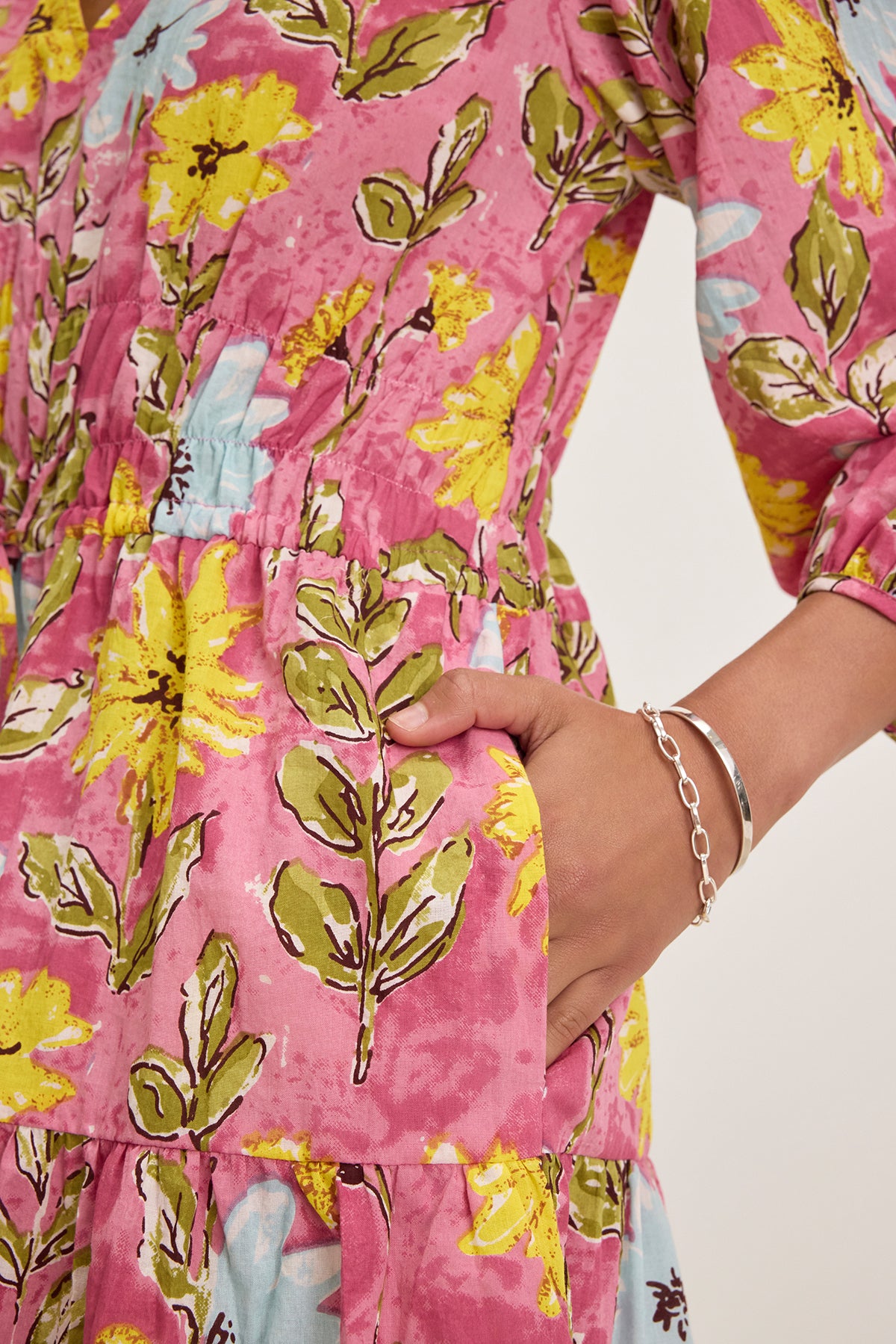   Wearing the SHARLENE DRESS by Velvet by Graham & Spencer, a person showcases a floral mini dress with yellow and green patterns on pink, featuring a smocked elastic waist, pockets, and v-neckline. They complete the look with a delicate silver bracelet on their wrist. 