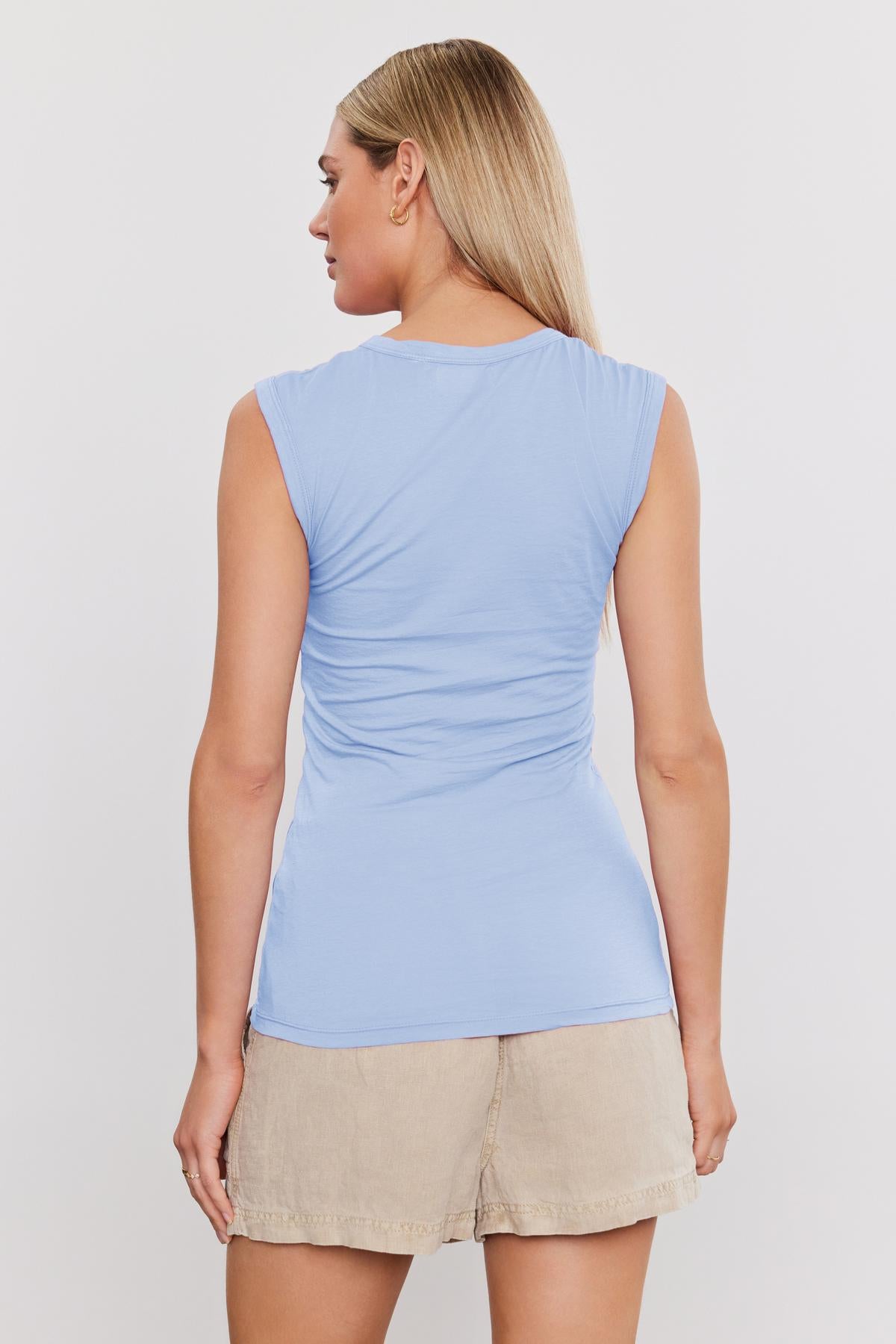   A person with long blonde hair is seen from the back, wearing a light blue Velvet by Graham & Spencer ESTINA TANK TOP and beige shorts. 