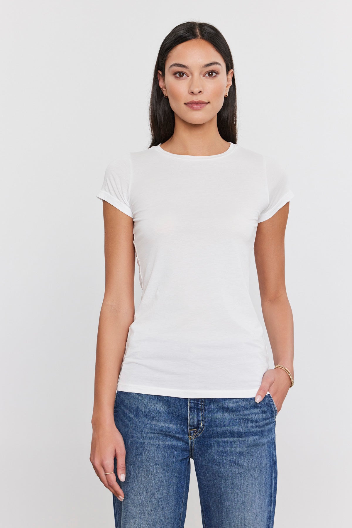   A woman with long dark hair is standing against a plain background, wearing a fitted crew neck white JEMMA TEE from Velvet by Graham & Spencer and blue jeans. 