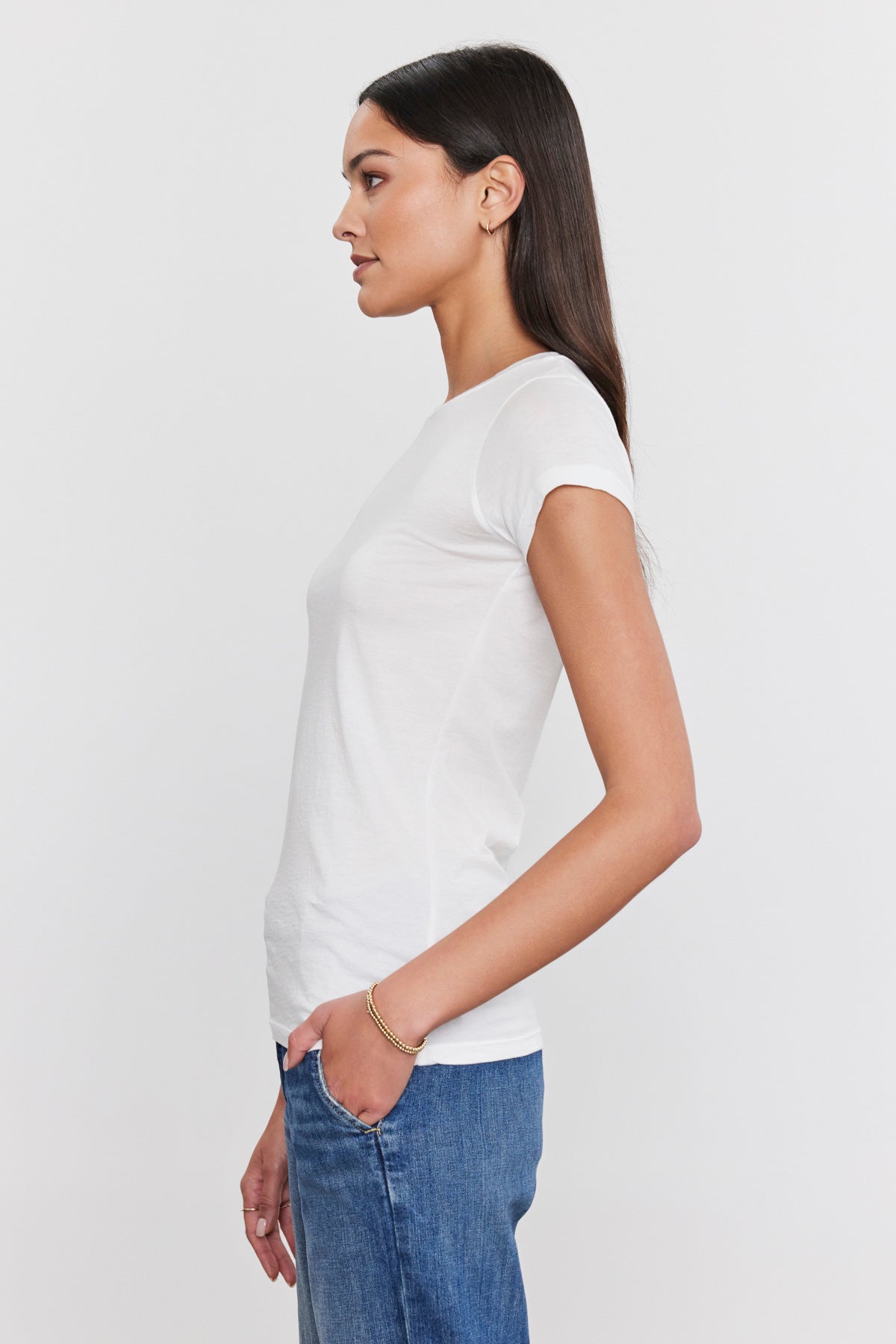 A woman with long, straight hair stands in profile, wearing a JEMMA TEE by Velvet by Graham & Spencer and blue jeans with her hand in her pocket.-35586101313729