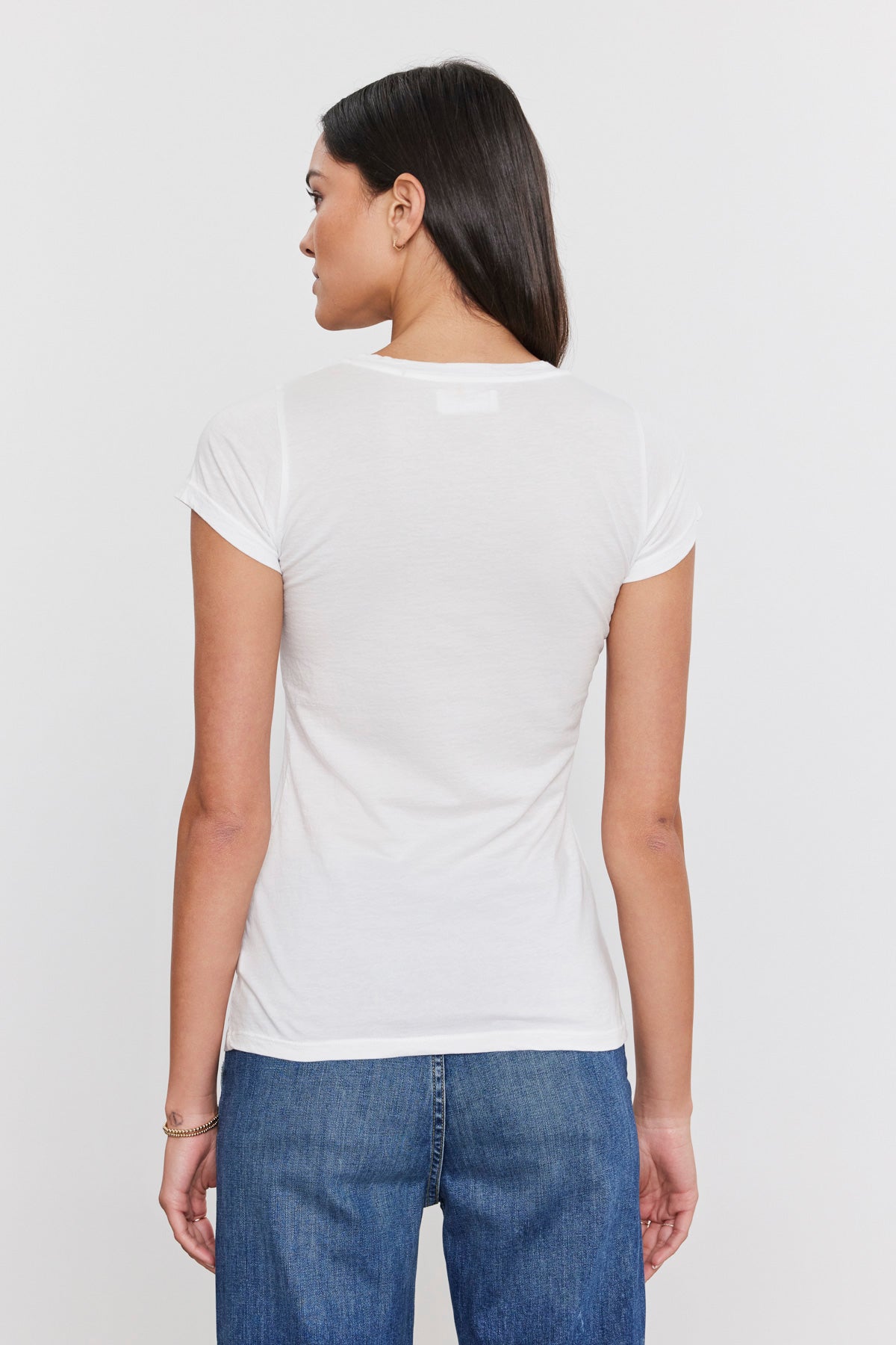   A person is standing facing away, wearing a fitted crew neck plain white short-sleeve Velvet by Graham & Spencer JEMMA TEE and blue jeans against a white background. 