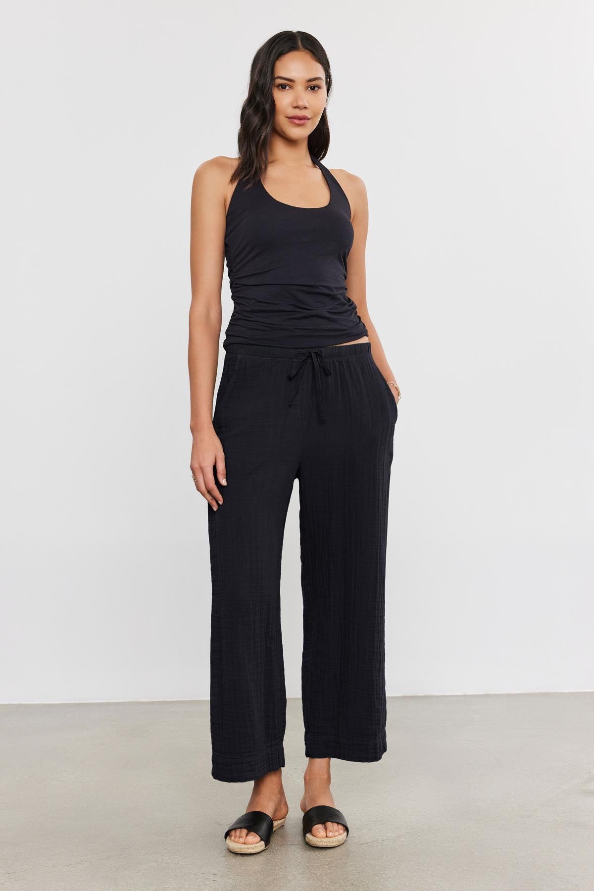   A woman stands against a plain background, wearing a sleeveless black top, FRANNY COTTON GAUZE PANT by Velvet by Graham & Spencer with slash pockets and a relaxed leg, and black sandals. She has long, dark hair and her hands are in her pockets. 