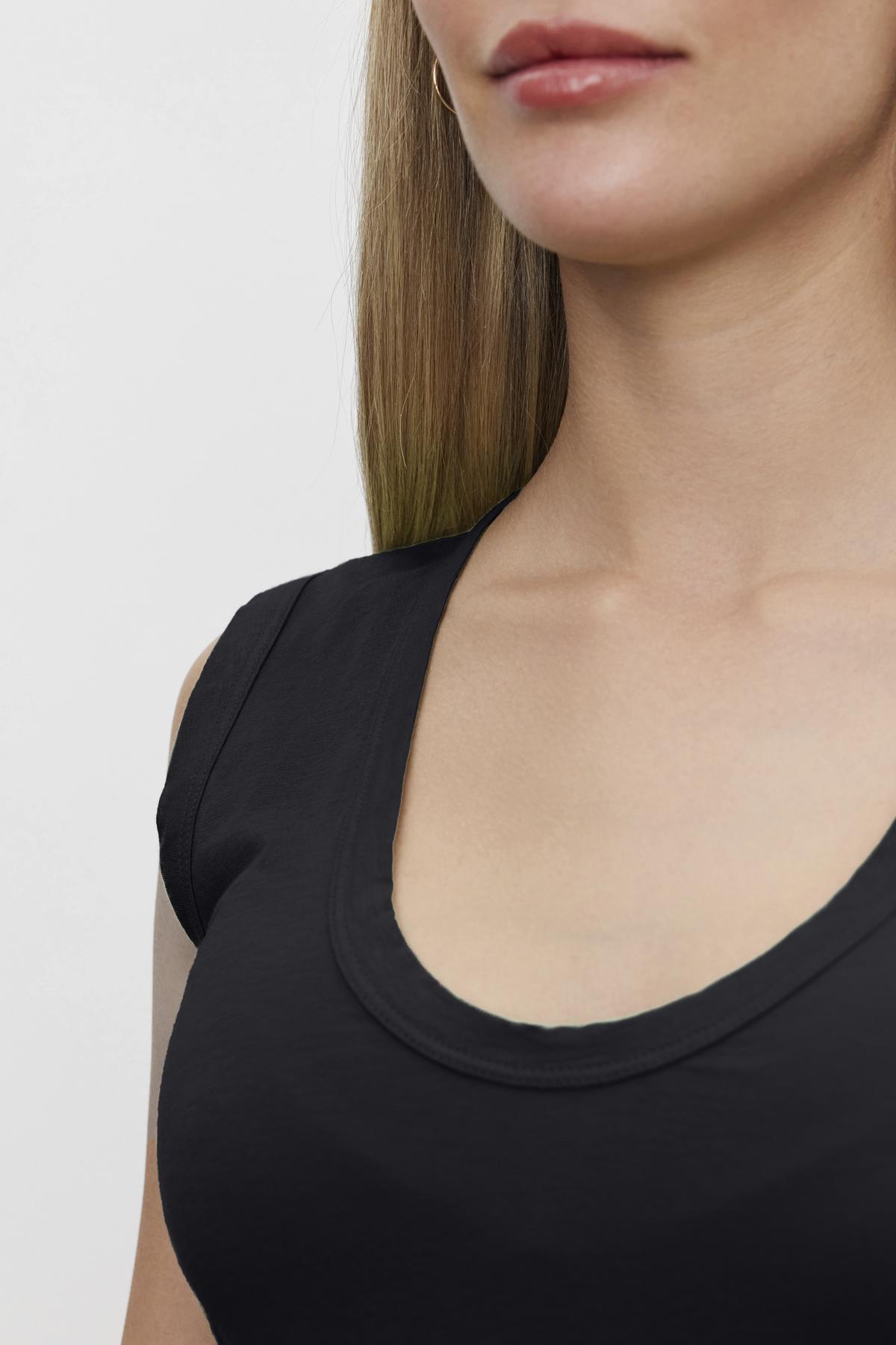 Close-up of a woman wearing a black ESTINA TANK TOP by Velvet by Graham & Spencer, showcasing the neckline and part of her face, with straight, light brunette hair against a plain background.-36266808443073