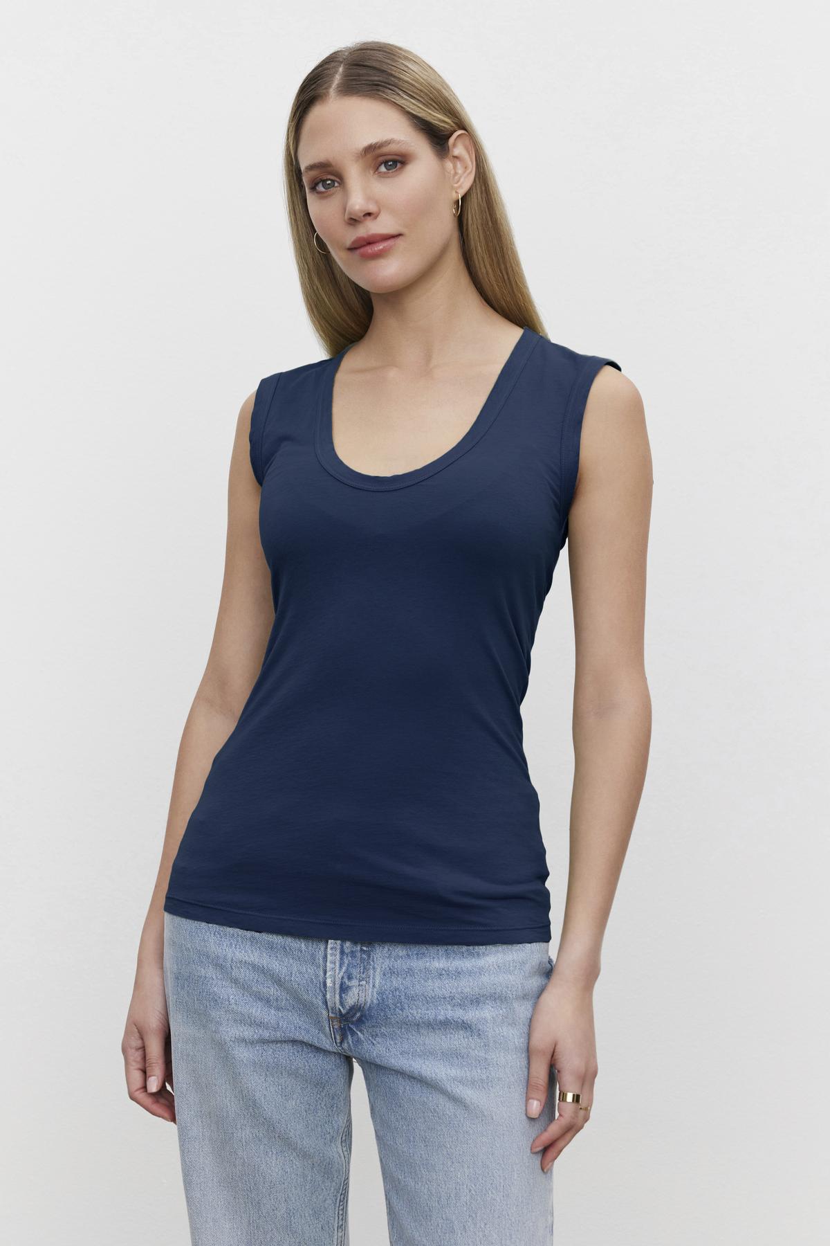   A woman with long hair is wearing a navy blue low-scoop-neck sleeveless Velvet by Graham & Spencer ESTINA TANK TOP and light blue jeans, standing against a plain white background. 