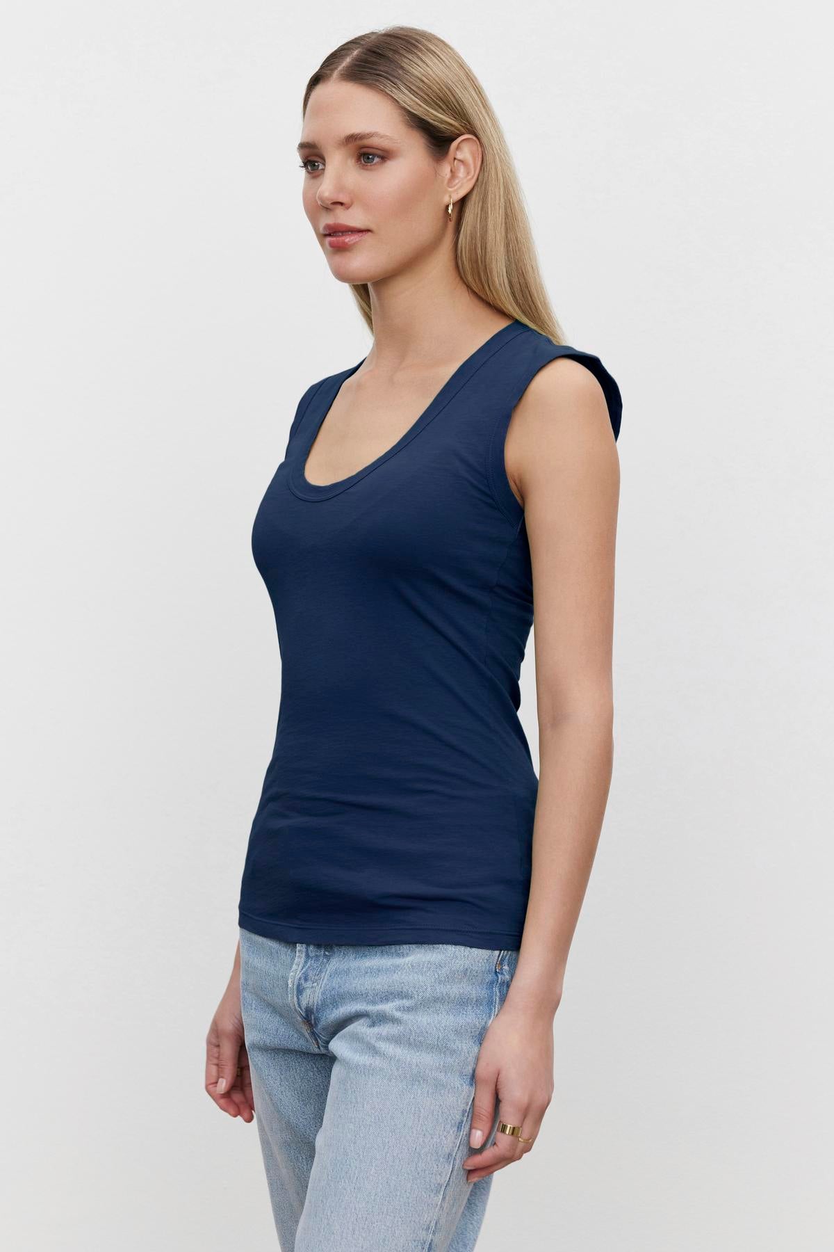 A woman with straight blond hair is wearing a navy blue, low-scoop-neck ESTINA TANK TOP by Velvet by Graham & Spencer and light blue jeans. She is standing against a plain white background, looking off to the side.-36273869619393