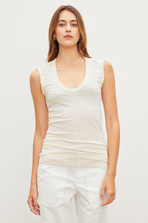 A woman stands facing the camera, wearing a sleeveless low-scoop-neck white top and white pants against a plain light background. The ESTINA TANK TOP by Velvet by Graham & Spencer exudes an effortless style, capturing a soft gauzy whisper that complements her relaxed demeanor.