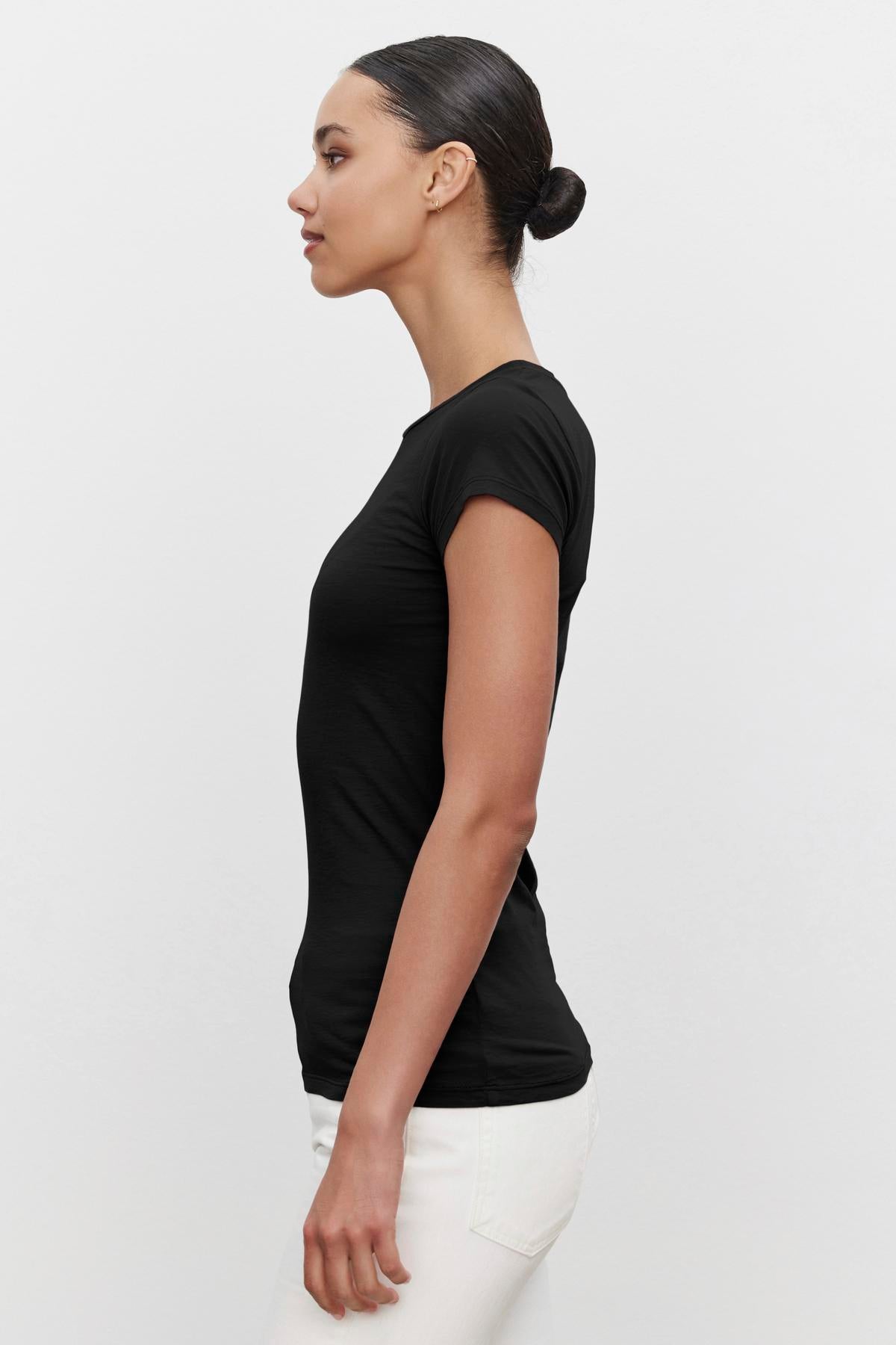   Profile view of a person with tied-back hair wearing the ultra-soft gauzy whisper JEMMA TEE in black by Velvet by Graham & Spencer and white pants against a plain background. 