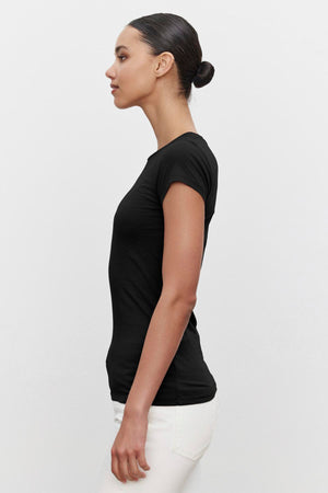 Profile view of a person with tied-back hair wearing the ultra-soft gauzy whisper JEMMA TEE in black by Velvet by Graham & Spencer and white pants against a plain background.