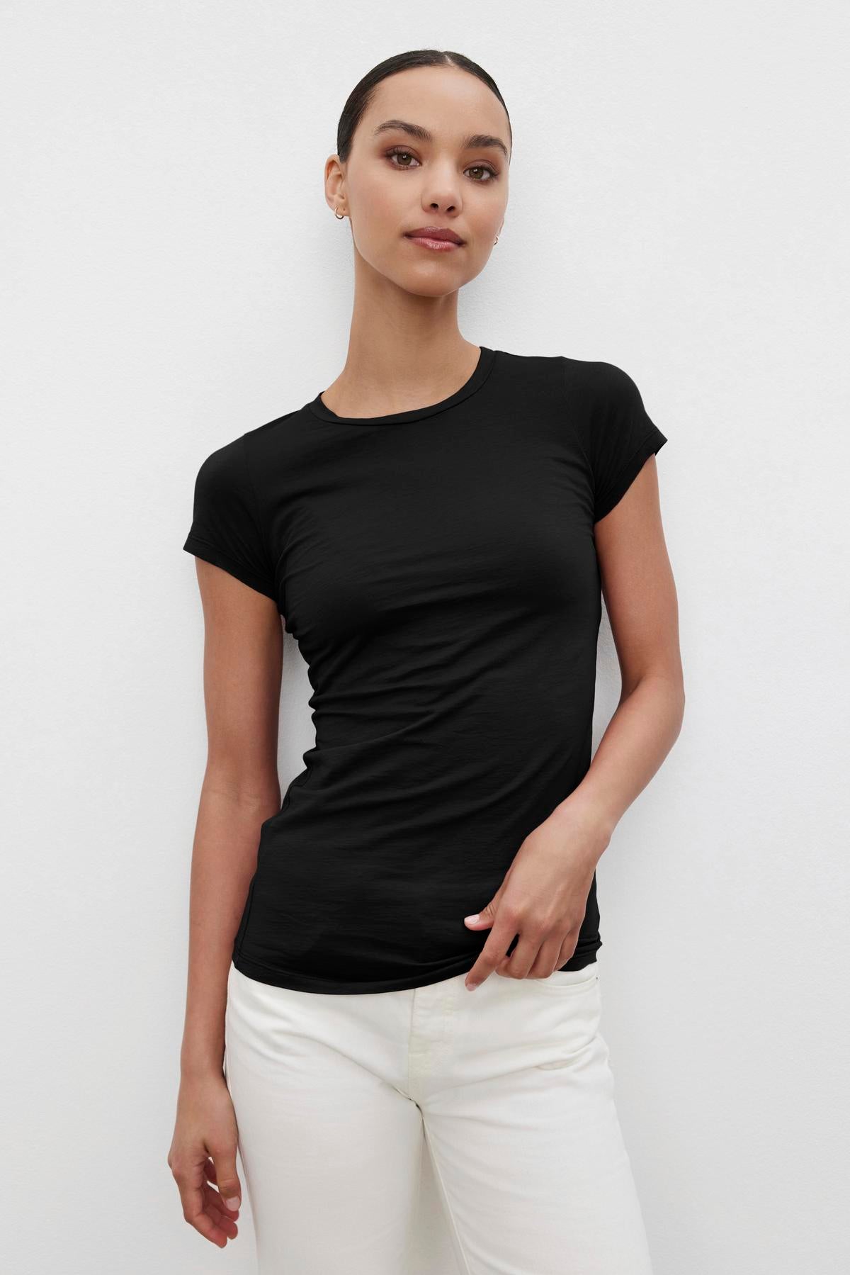   A person wearing the JEMMA TEE in a gauzy whisper black crew neck by Velvet by Graham & Spencer, paired with white pants, stands against a plain white background, facing the camera with a neutral expression. 