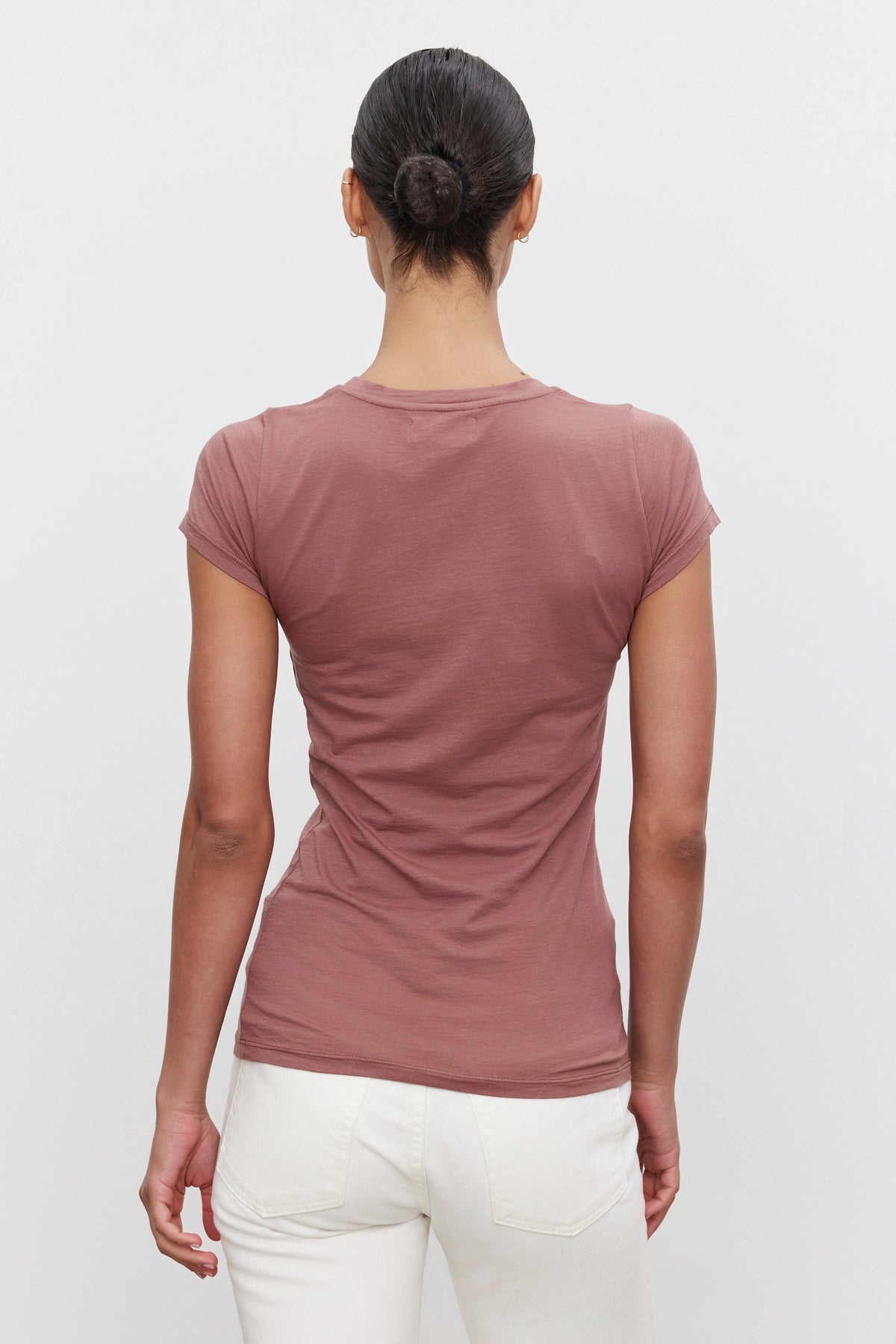 A person with dark hair in a bun is standing with their back to the camera. They are wearing an ultra-soft gauzy whisper short-sleeve, mauve-colored JEMMA TEE by Velvet by Graham & Spencer and white pants.-37386168762561