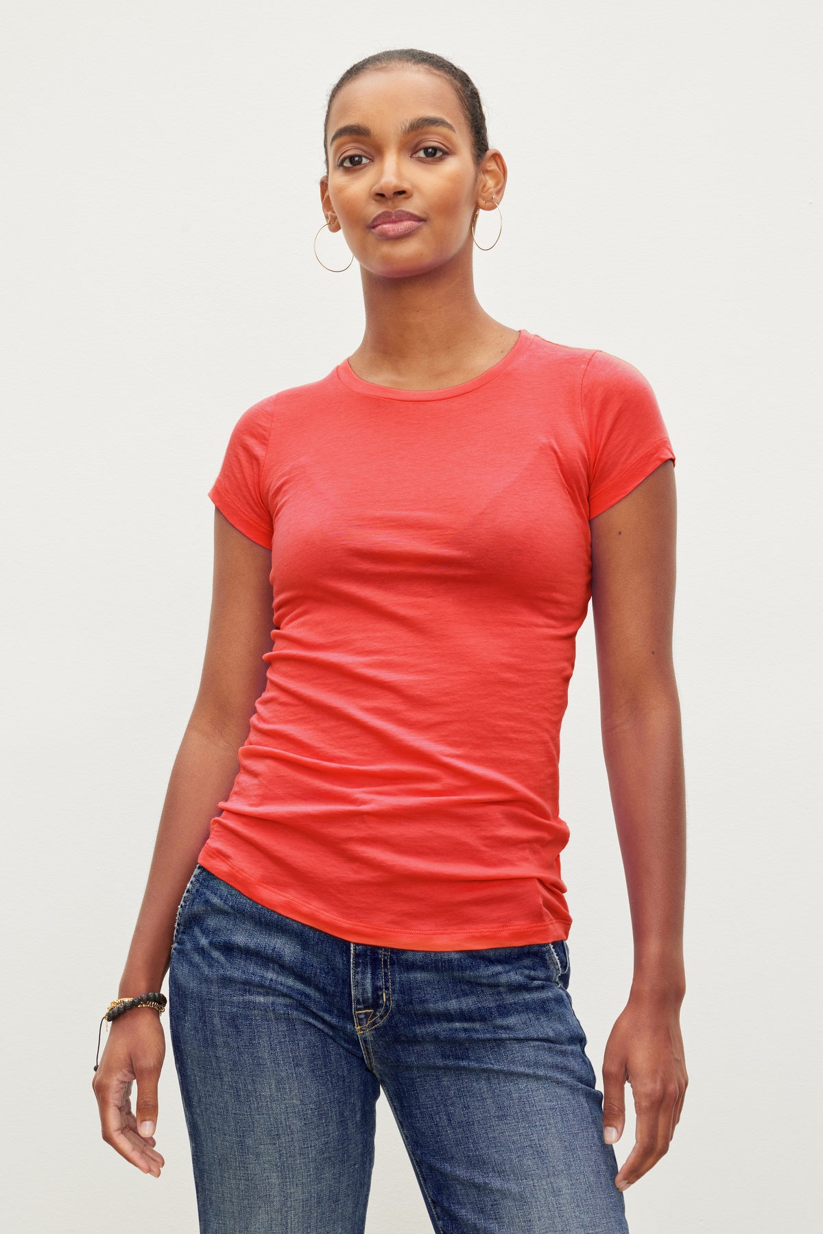   A person with short hair wearing a fitted crew neck red JEMMA TEE by Velvet by Graham & Spencer and blue jeans poses against a plain background. 