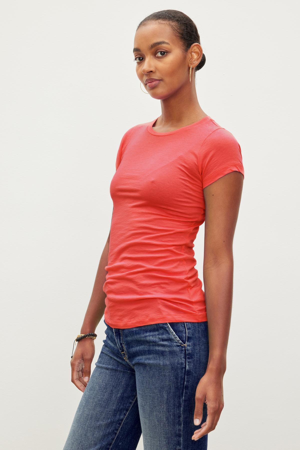 A person wearing a fitted crew neck red short-sleeve JEMMA TEE by Velvet by Graham & Spencer and blue jeans stands against a plain white background.-36533000208577