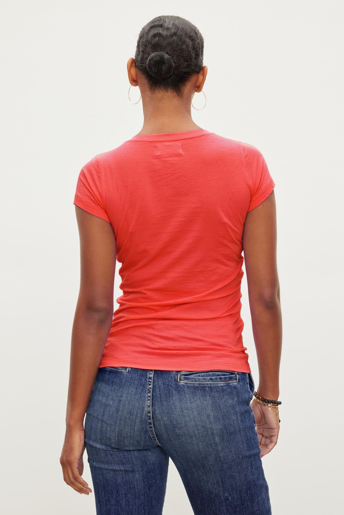   A person in a red JEMMA TEE by Velvet by Graham & Spencer and blue jeans is standing, facing away, with hair in a bun and wearing hoop earrings. 