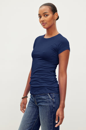 A person with short hair wears a fitted crew neck navy blue JEMMA TEE by Velvet by Graham & Spencer and blue jeans, standing against a plain background.