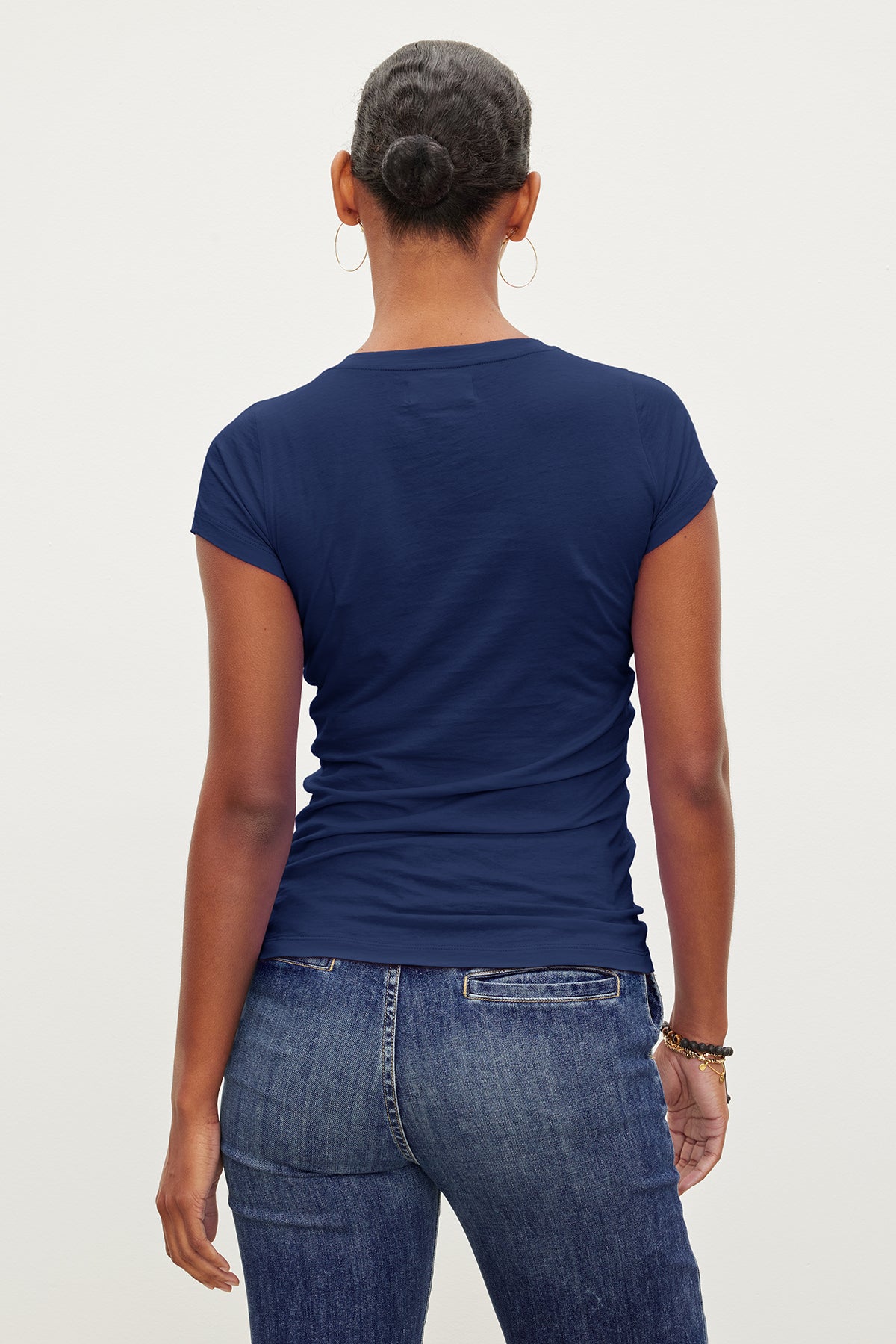   A person is shown from the back, wearing a JEMMA TEE by Velvet by Graham & Spencer, which is a fitted crew neck navy blue short-sleeved shirt, and blue jeans. Their hair is neatly styled in a bun. 