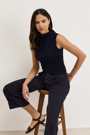 A woman in a black sleeveless top and Velvet by Graham & Spencer's VERA CORDUROY PANT sits on a wooden stool against a plain wall.