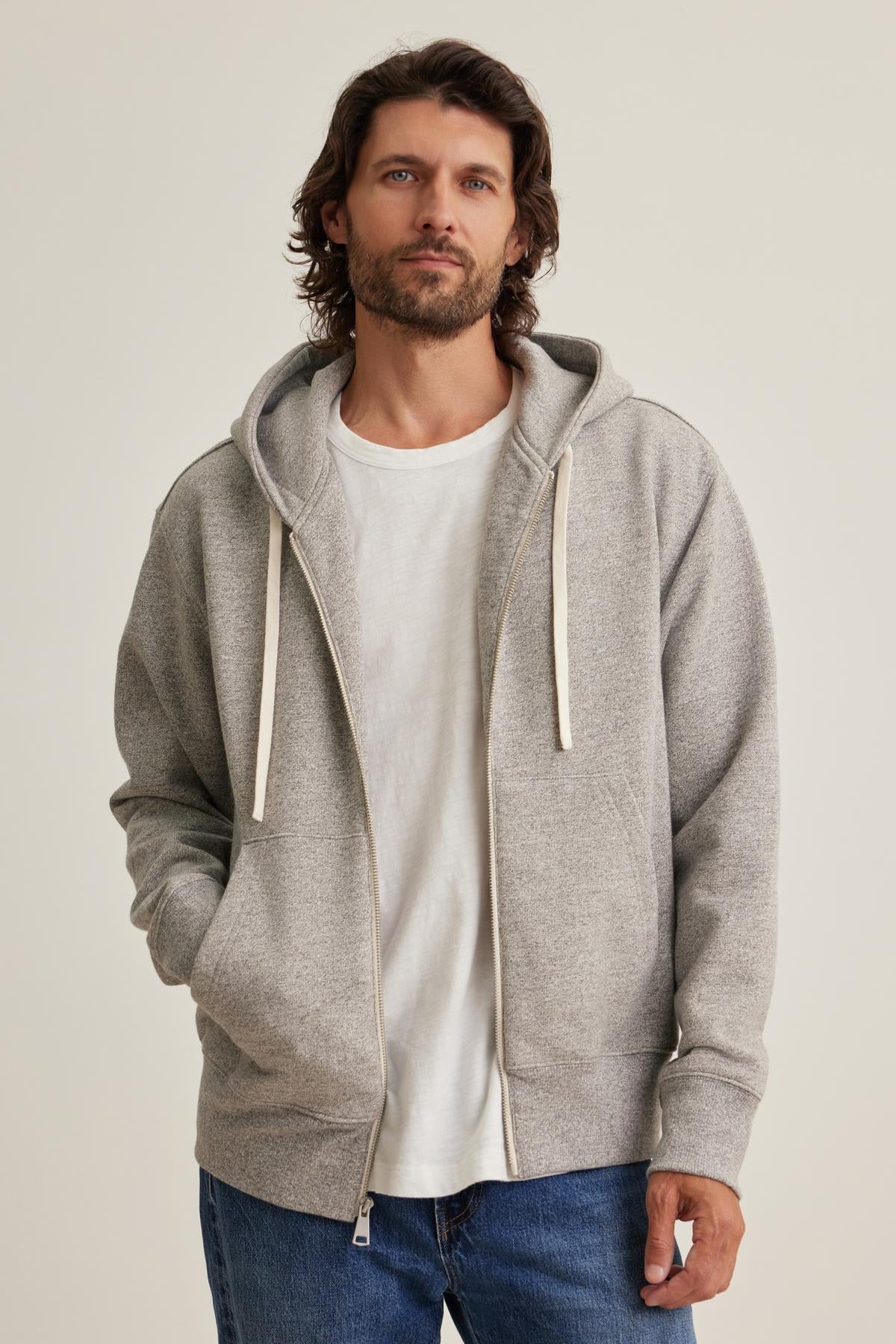   A man wears the DRACO HOODIE by Velvet by Graham & Spencer, a gray zip-up over a white t-shirt and blue jeans, standing with hands in pockets against a plain background. 