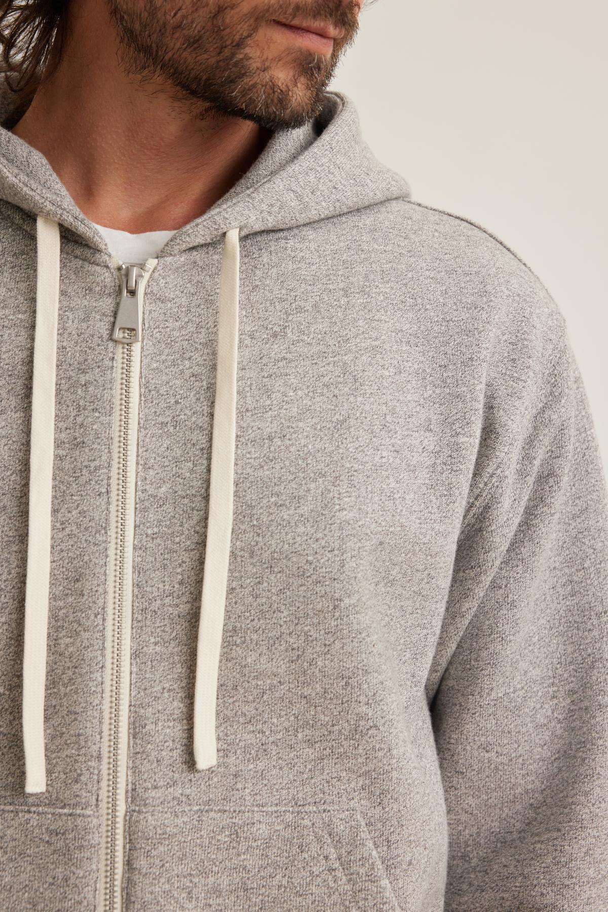 A man wears the DRACO HOODIE by Velvet by Graham & Spencer, a gray zip-up hoodie featuring white drawstrings, with his partially visible head and beard set against a light background.-38643384221889