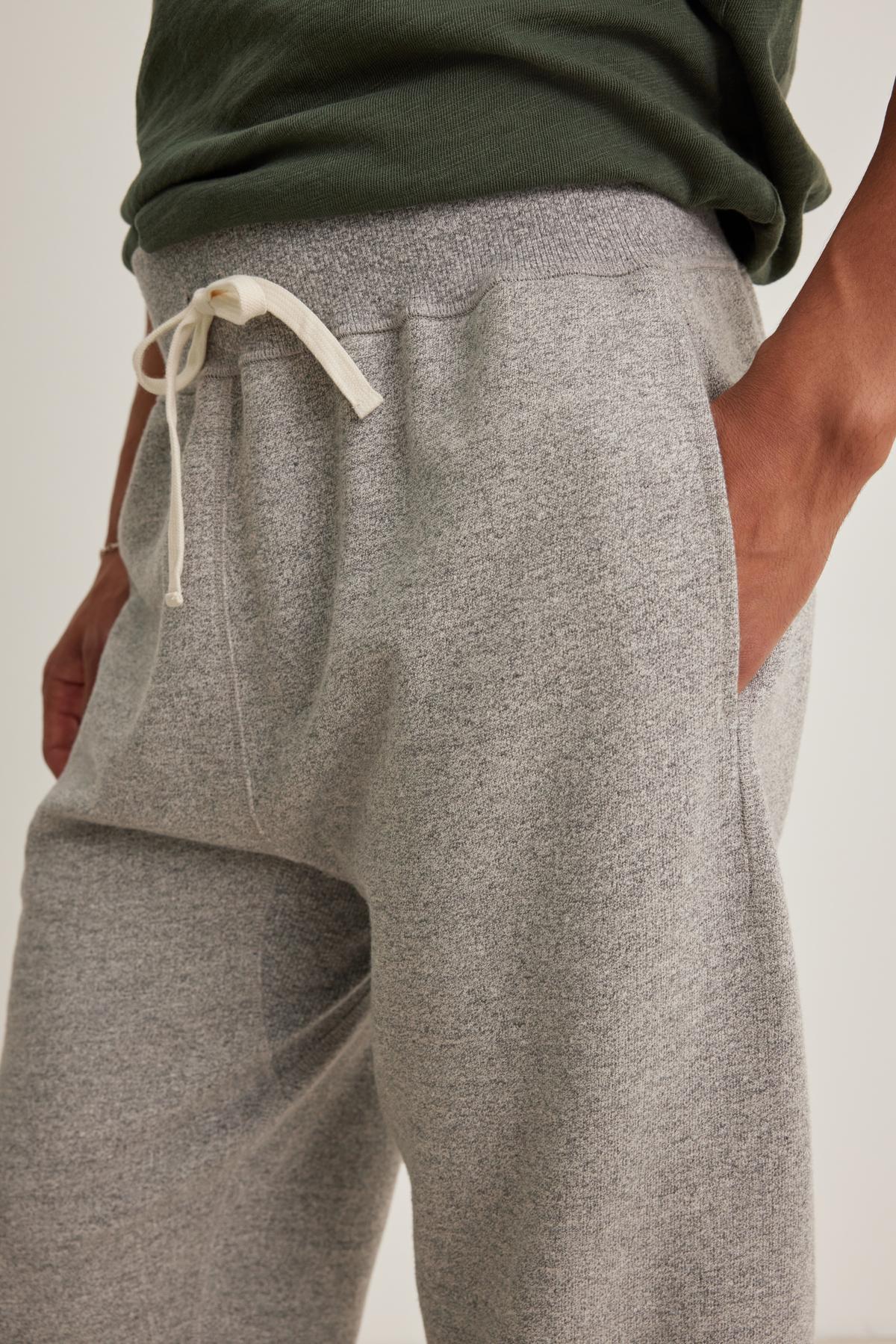   A close-up shows a person wearing Velvet by Graham & Spencer's KEELAN JOGGER, featuring grey sweatpants with a white drawstring and pockets, paired with a dark green shirt. 