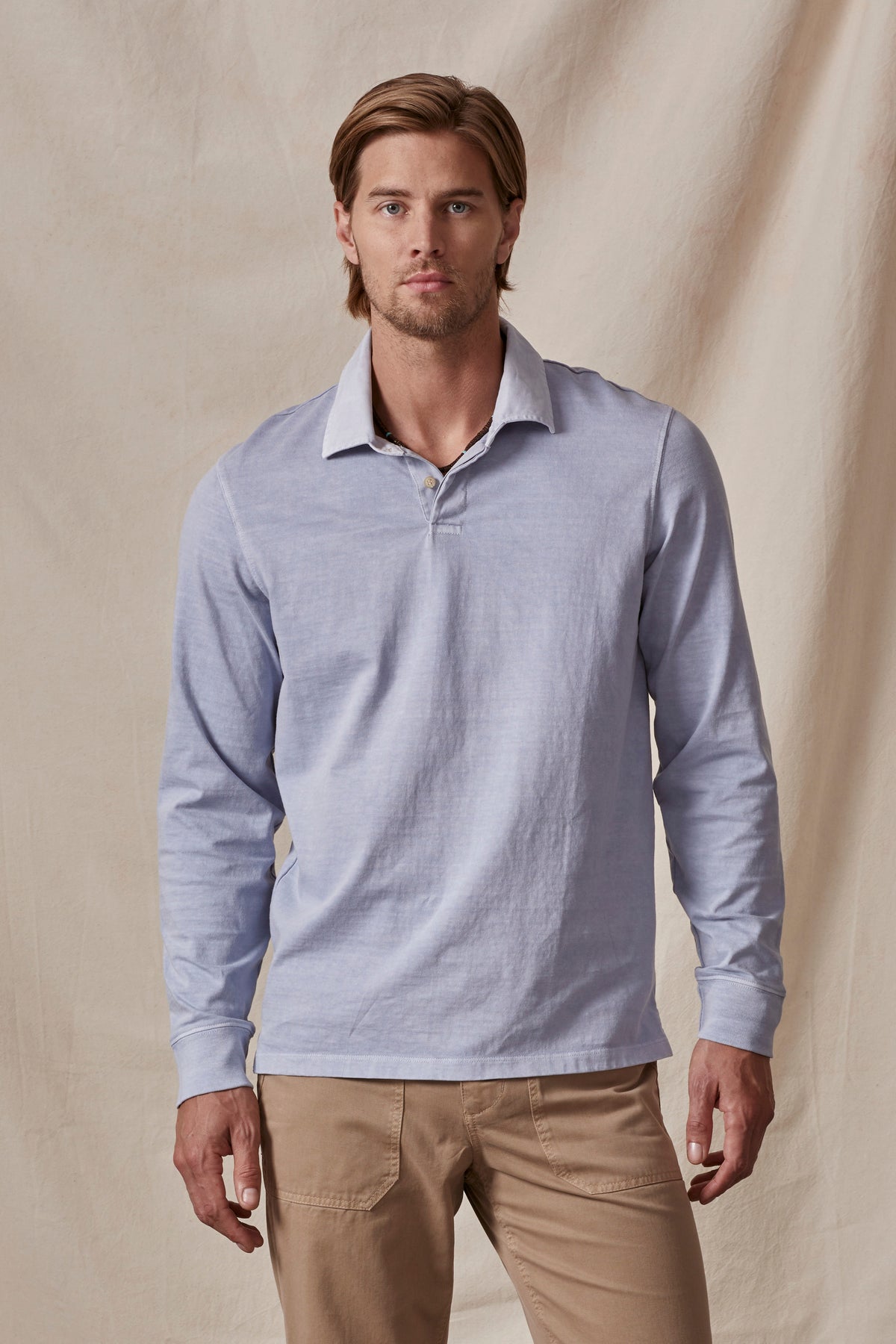   A man wearing a light blue long-sleeve BALTHAZAR POLO by Velvet by Graham & Spencer and beige pants stands in front of a beige fabric backdrop. 