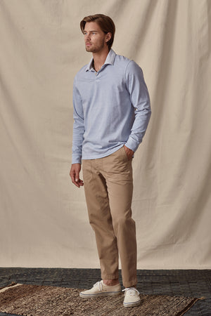 A man wearing a relaxed fit light blue long-sleeve BALTHAZAR POLO by Velvet by Graham & Spencer, beige pants, and white sneakers stands against a beige backdrop.