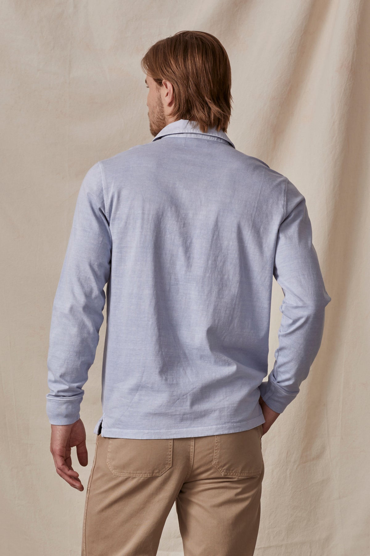   A person with light brown hair is facing away, wearing a light blue long-sleeve Velvet by Graham & Spencer BALTHAZAR POLO and beige pants. The background is a beige fabric. 