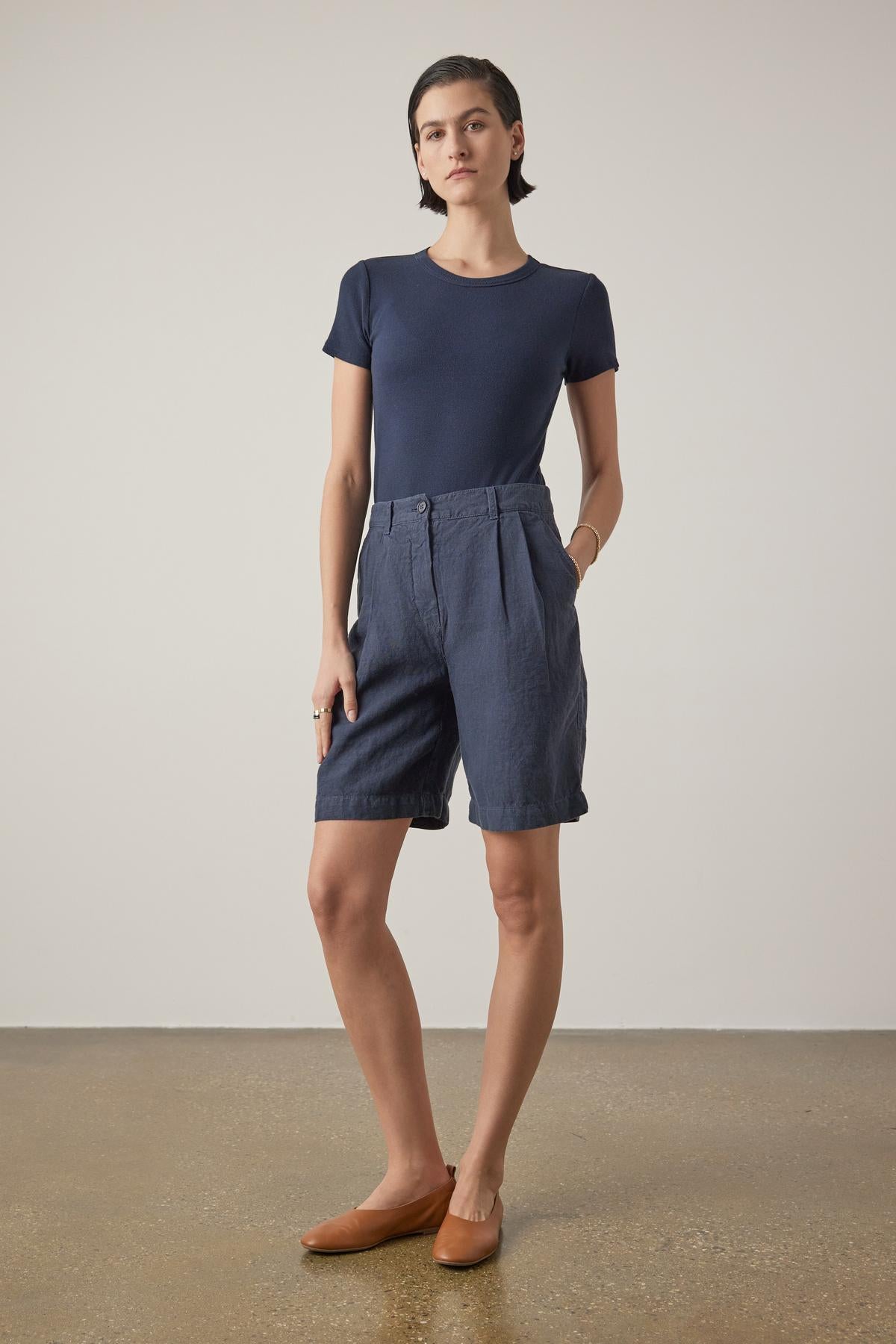   Person standing, wearing a navy blue t-shirt, Velvet by Jenny Graham's dark blue LARCHMONT HEAVY LINEN SHORT, and tan flats, showcasing warm-weather styling against a plain background. 