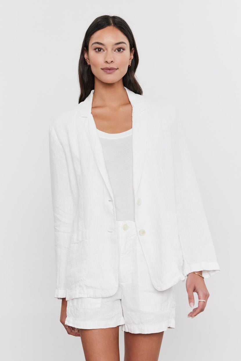 A woman in a spring suit consisting of a Velvet by Graham & Spencer LENNY HEAVY LINEN BLAZER and shorts set, paired with a white t-shirt, standing against a plain background.