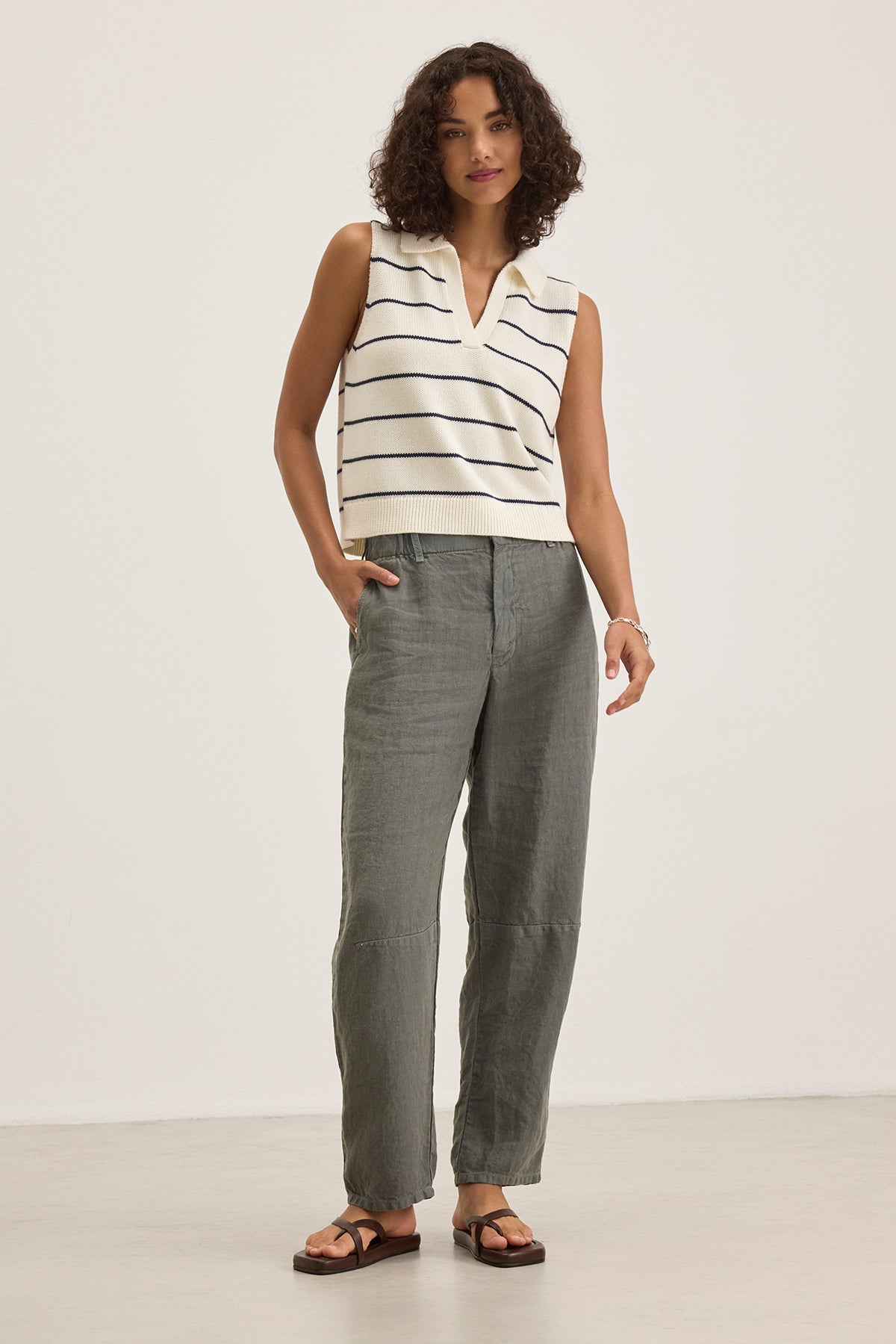   A person stands against a plain background wearing the Velvet by Graham & Spencer NAYLA Cotton Cashmere Sweater Vest over a sleeveless white striped top with gray pants and sandals, showcasing layered comfort and style. 