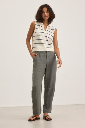 A person stands against a plain background wearing the Velvet by Graham & Spencer NAYLA Cotton Cashmere Sweater Vest over a sleeveless white striped top with gray pants and sandals, showcasing layered comfort and style.