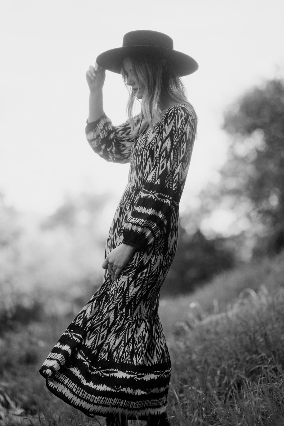   Black-and-white image of a woman wearing the Velvet by Graham & Spencer CATHERINE DRESS, featuring an ikat print, and a wide-brimmed hat, touching the brim of her hat as she stands outdoors with greenery in the background. 