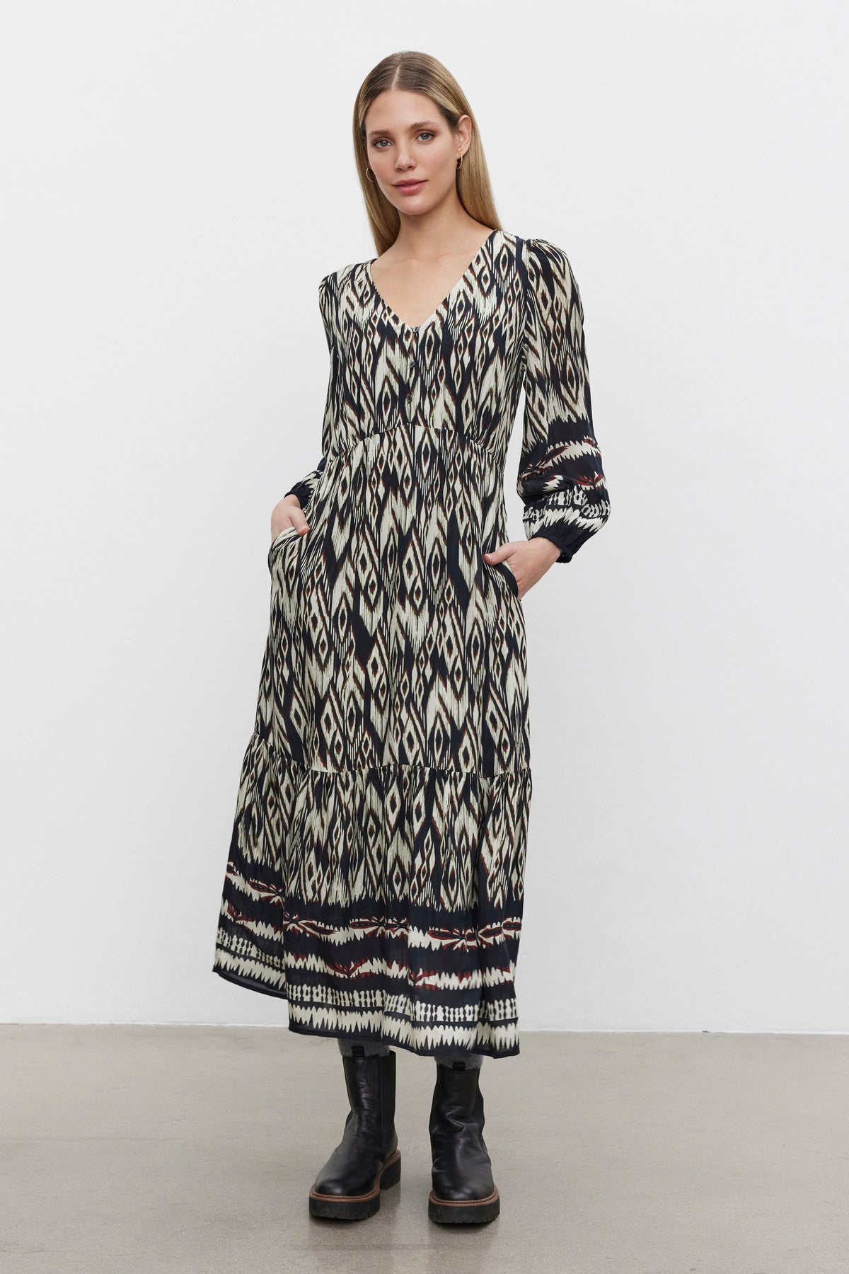   A woman stands against a white background wearing the CATHERINE DRESS by Velvet by Graham & Spencer; it features a long-sleeved, black-and-white patterned ikat print with pockets, paired with black platform boots. 