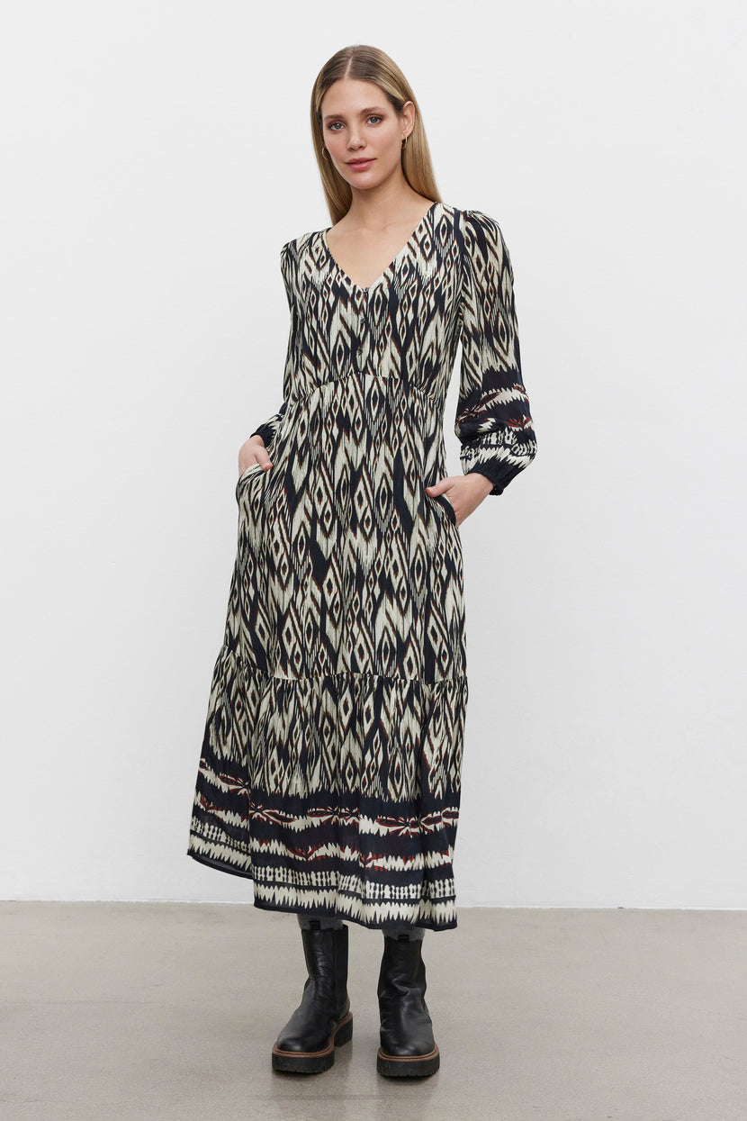 A woman stands against a white background wearing the CATHERINE DRESS by Velvet by Graham & Spencer; it features a long-sleeved, black-and-white patterned ikat print with pockets, paired with black platform boots.