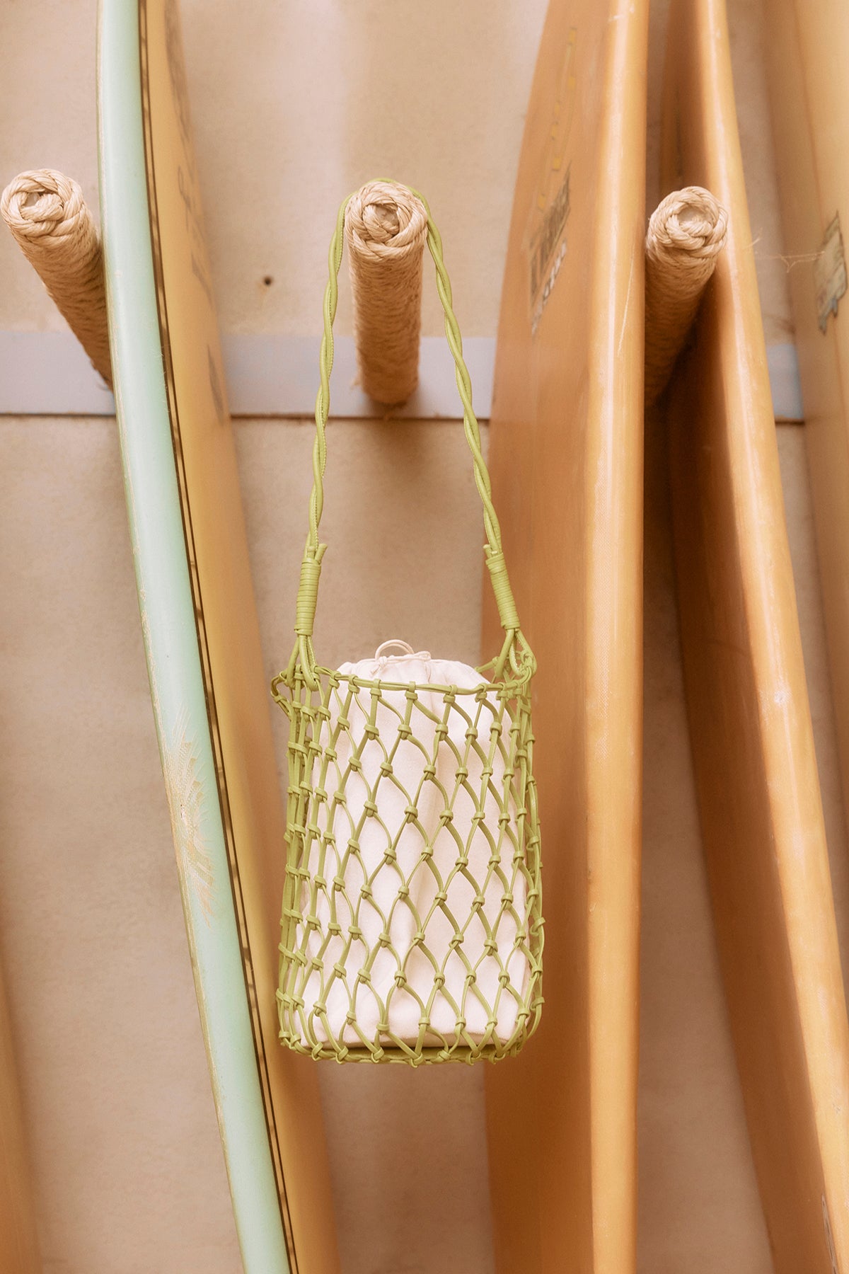   A green Velvet by Graham & Spencer mesh tote bag hanging from a wooden bamboo pole, with other bamboo poles in the background. 