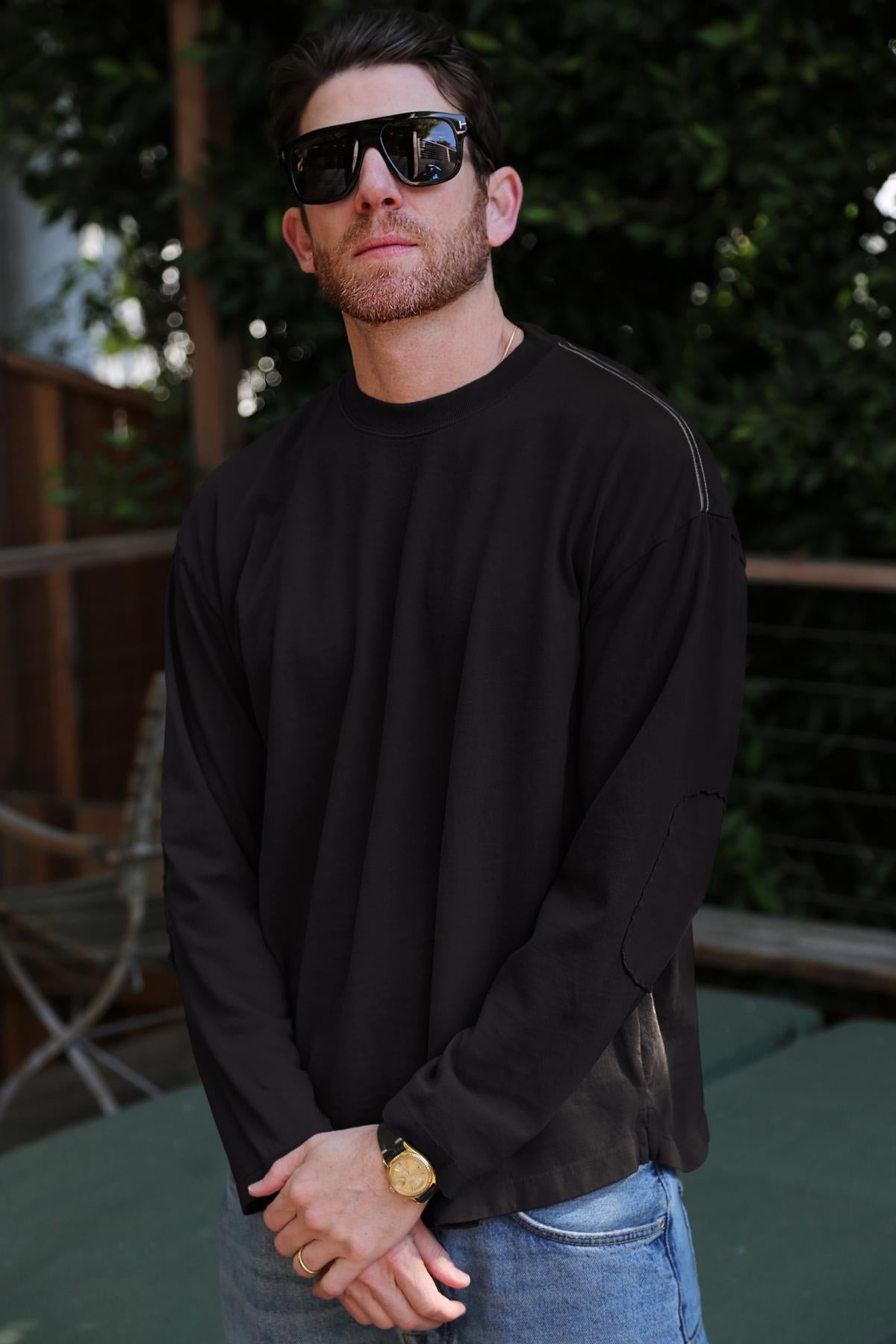   Wearing sunglasses, a SLATER TEE by Velvet by Graham & Spencer in a relaxed fit black long-sleeve made of luxurious Pima cotton jersey, and jeans, a man stands outdoors amidst greenery and a wooden railing. 