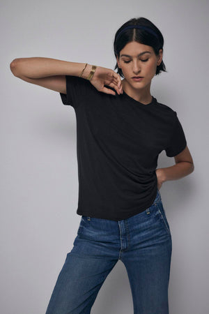 A person with short dark hair wearing a classic black Velvet by Jenny Graham SOLANA TEE and tailored blue jeans poses against a plain background.