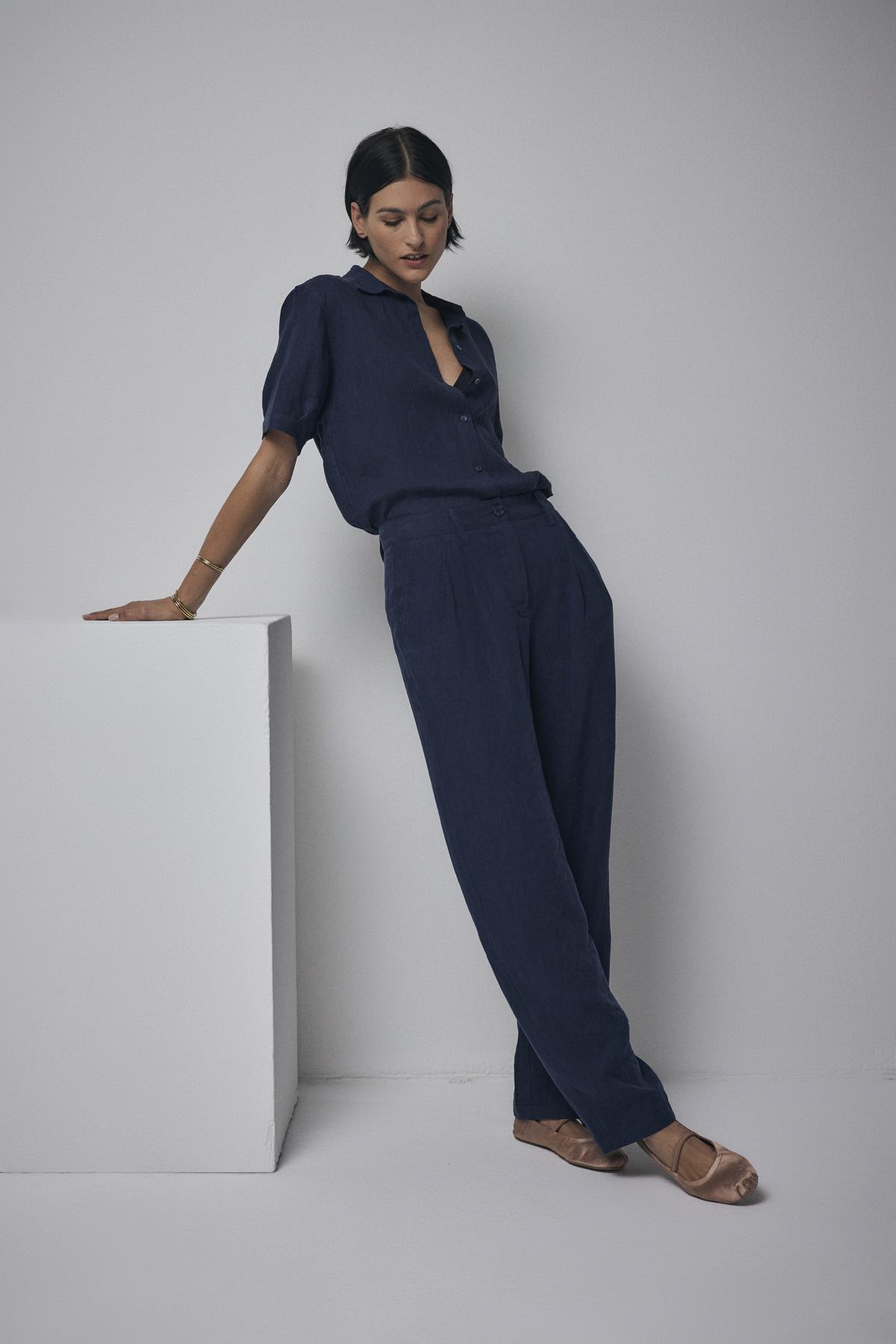   A woman in a CLAREMONT LINEN SHIRT from Velvet by Jenny Graham leans on a white cube in a minimalist setting. 