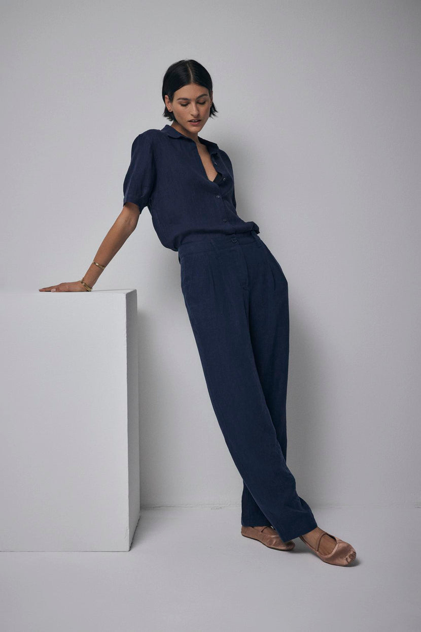 A woman in a CLAREMONT LINEN SHIRT from Velvet by Jenny Graham leans on a white cube in a minimalist setting.