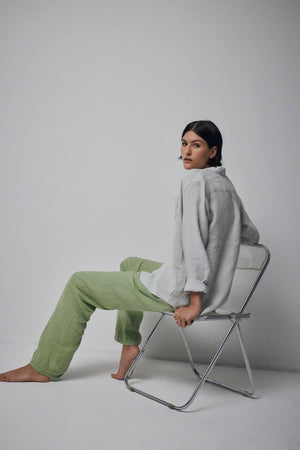 A person with long dark hair, dressed in a relaxed silhouette featuring a light-colored MULHOLLAND LINEN SHIRT by Velvet by Jenny Graham and green pants, sits barefoot on a folding chair against a plain backdrop.