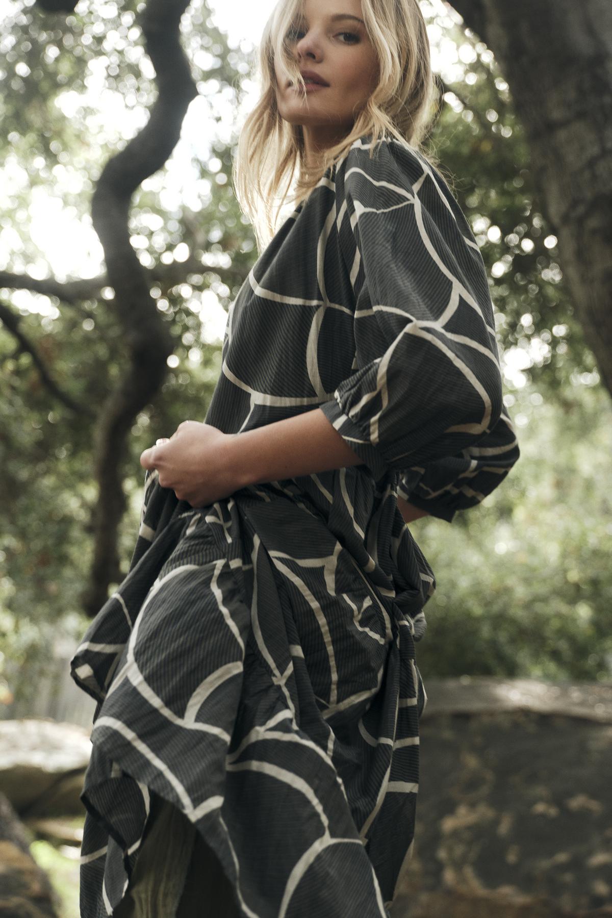   A woman stands outdoors, wearing the JOHANNA PRINTED SILK COTTON VOILE DRESS by Velvet by Graham & Spencer. The black and white patterned design with a bold print complements her stylish look, as trees and plants create a beautiful backdrop. 