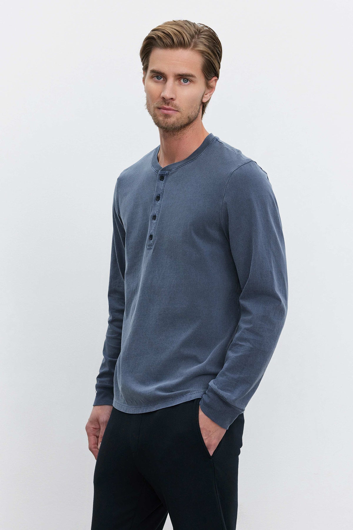 A person with light brown hair, wearing the REMI HENLEY by Velvet by Graham & Spencer—a medium-weight cotton knit blue long-sleeve Henley shirt—and black pants, stands against a plain white background with one hand in a pocket and a neutral expression.-37643196235969