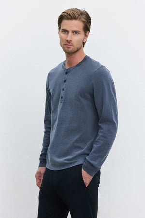 A person with light brown hair, wearing the REMI HENLEY by Velvet by Graham & Spencer—a medium-weight cotton knit blue long-sleeve Henley shirt—and black pants, stands against a plain white background with one hand in a pocket and a neutral expression.