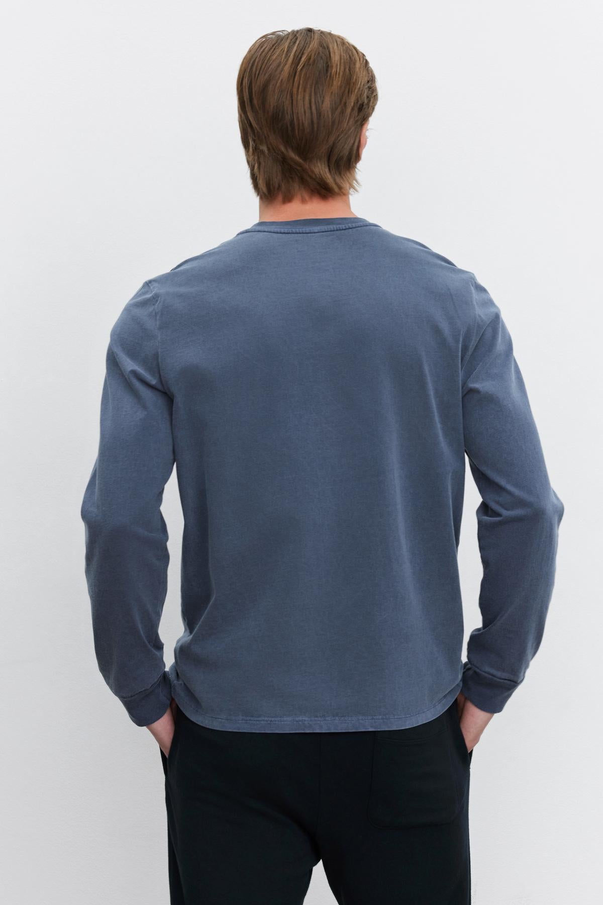   A person with light brown hair is seen from the back, wearing the Velvet by Graham & Spencer REMI HENLEY, a long-sleeved faded blue shirt with a reinforced button placket, along with black pants, standing against a plain white background. 