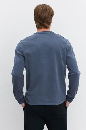 A person with light brown hair is seen from the back, wearing the Velvet by Graham & Spencer REMI HENLEY, a long-sleeved faded blue shirt with a reinforced button placket, along with black pants, standing against a plain white background.