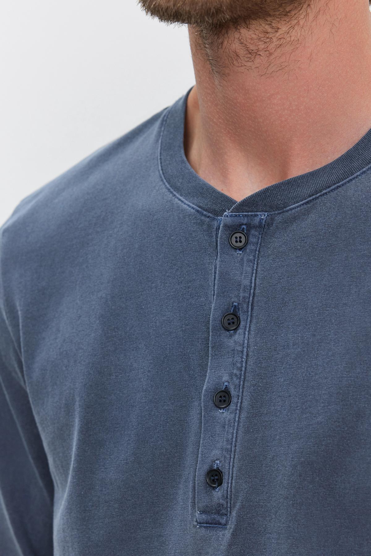   A close-up of a person wearing the REMI HENLEY by Velvet by Graham & Spencer, featuring a blue, long-sleeved shirt with a buttoned henley collar. The medium-weight cotton knit highlights the shirt's texture and meticulous button detail, complemented by a reinforced button placket for enhanced durability. 