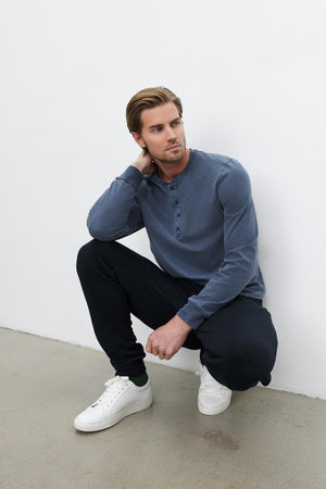 A man with light brown hair sits on the ground in a white room, wearing the REMI HENLEY by Velvet by Graham & Spencer, an indigo medium-weight cotton knit henley shirt with a reinforced button placket, paired with black pants and white sneakers. One hand rests on his knee while the other is positioned behind his head.